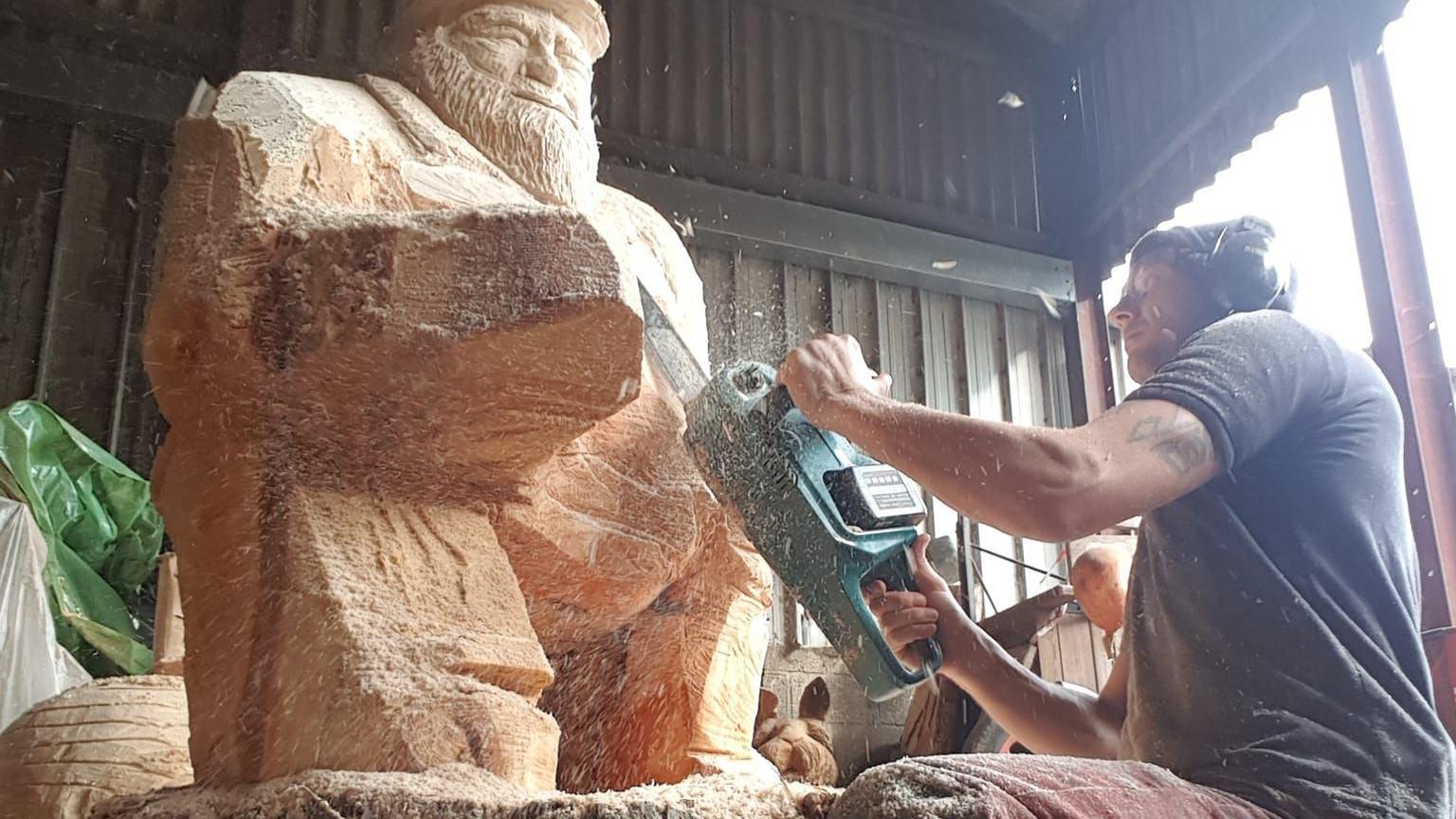 Graham Derham using a chainsaw, wearing ear defenders and a grey T-shirt, carving wood into a sculpture of a bearded man inside a barn.