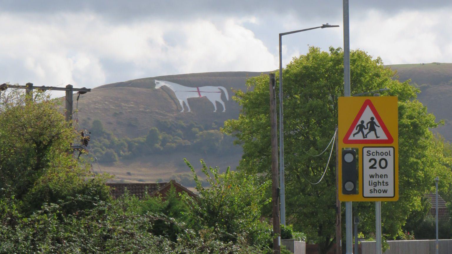 A large white horse carved into the side of a hill, with red fabric draped over it in a cross. In the foreground there are road signs and lampposts. 