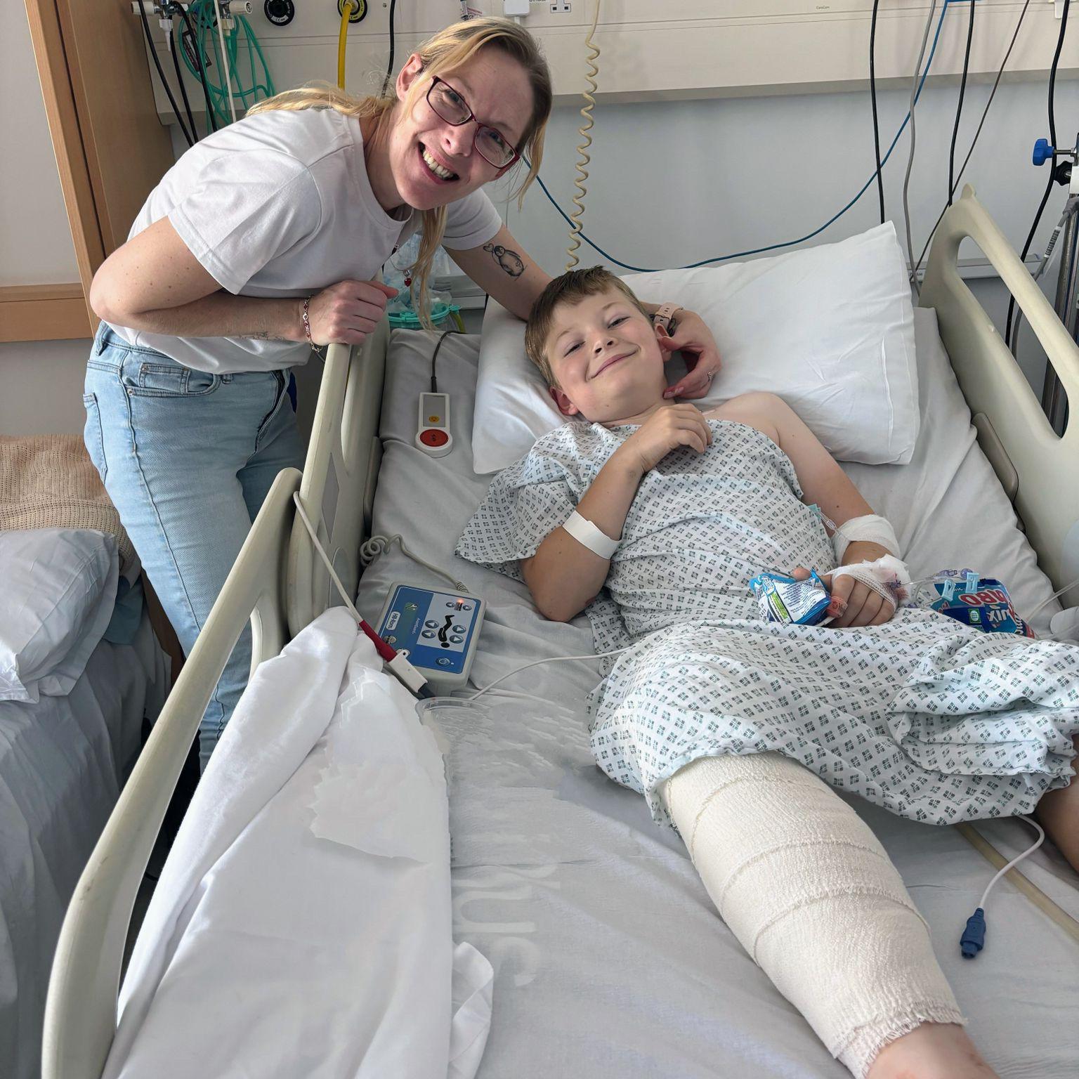 Jo Newman, who has blonde hair and glasses, with her son at Cardiff's Noah's Ark Children's Hospital. He is lying in a bed with a bandaged leg in a hospital gown
