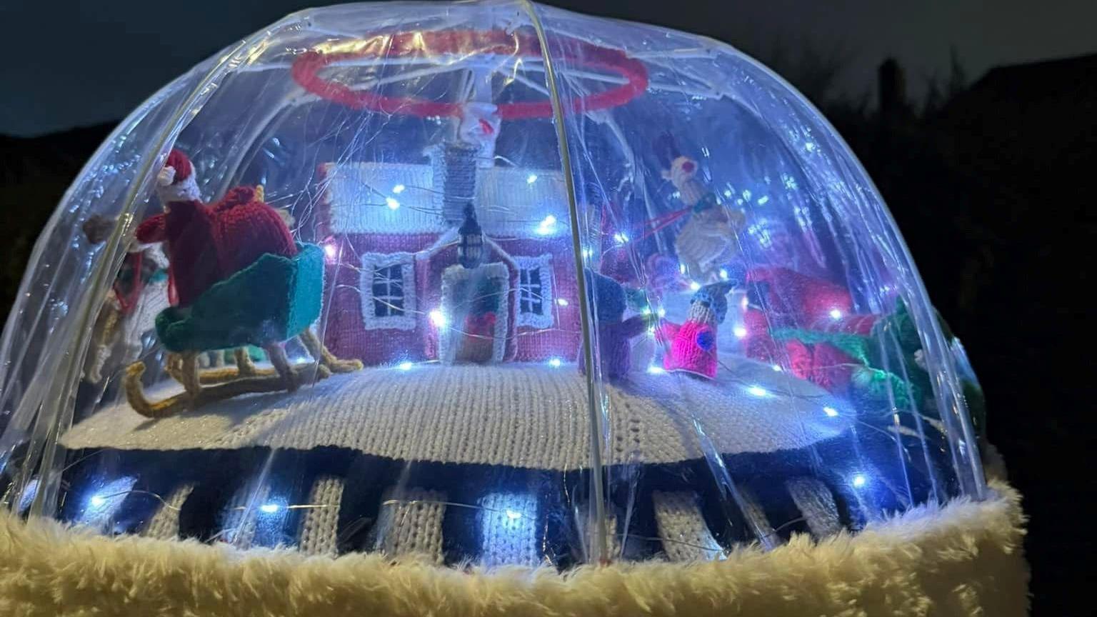 A scene protected by a clear plastic dome includes a knitted house with pink walls and a snowy, white roof, lit up for Christmas. Santa can be seen to one side, on his sleigh delivering presents. The whole display is lit up with white fairy lights.