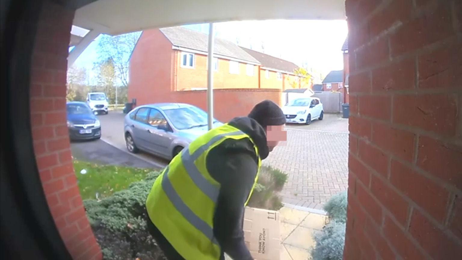 A picture taken from a doorbell camera showing a man wearing a high-viz jacket and a black hat. He has just picked up a parcel and is about to walk away.