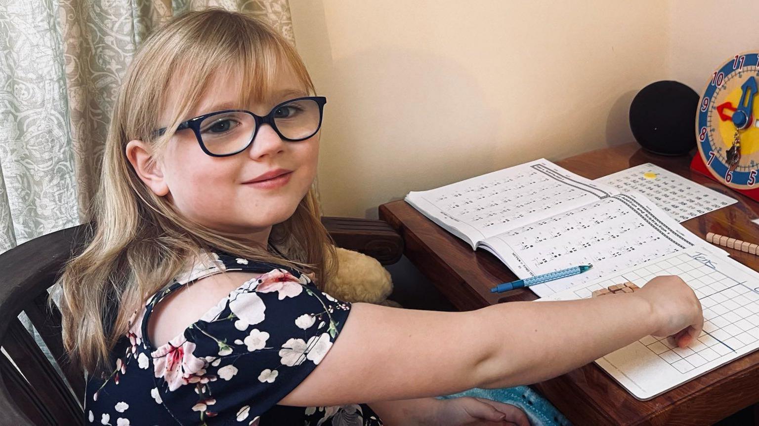 Blonde girl with glasses sits at a desk, looking at the camera and smiling. She has a maths textbook open and a whiteboard beside it. She has wooden blocks on the board and there is a toy clock behind it with the time reading 8 o'clock. 