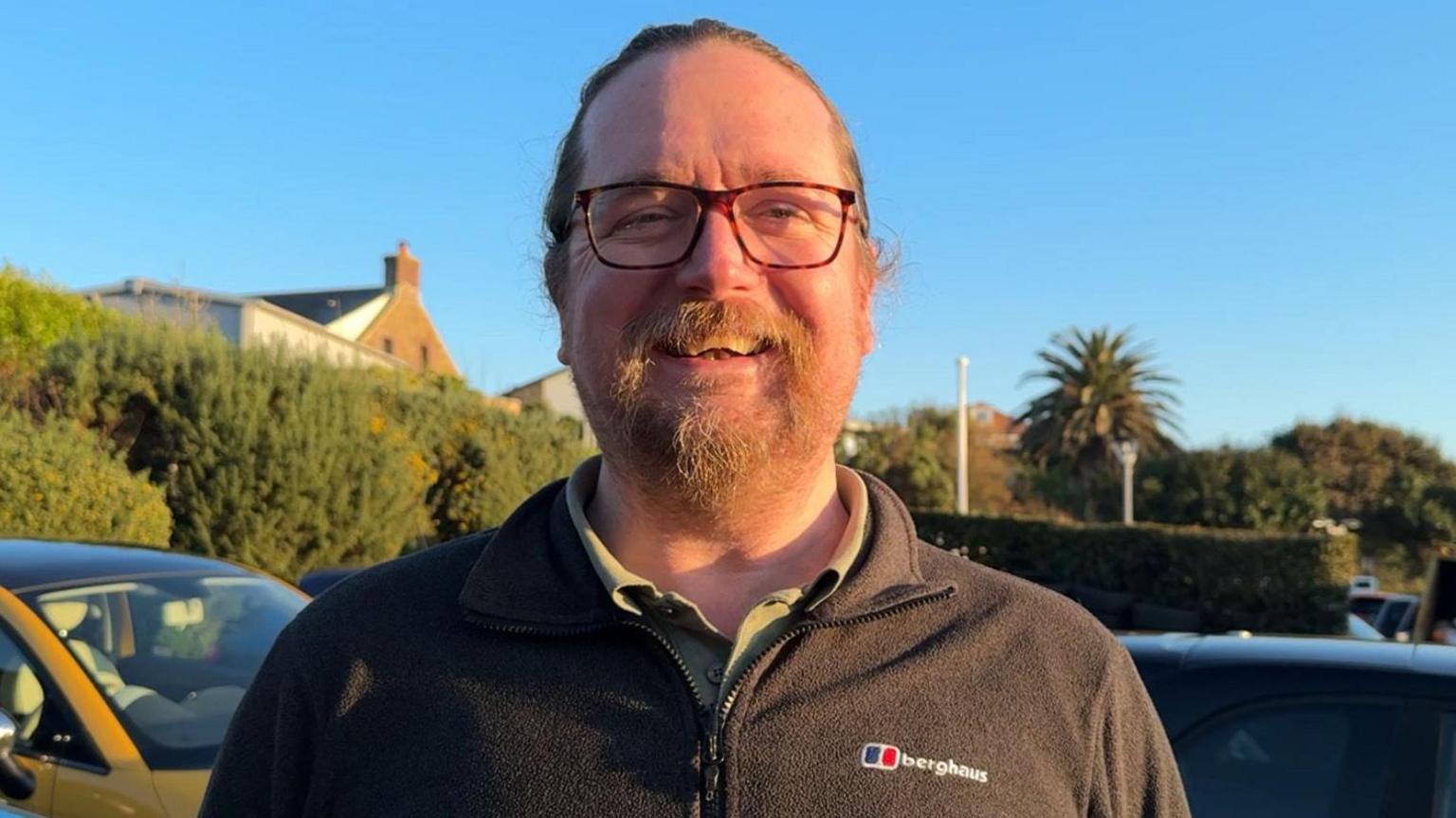 Olly Brock smiles on a sunny day while wearing a black Berghaus fleece and standing in front of several parked cars. He has glasses on and a large goatee beard.