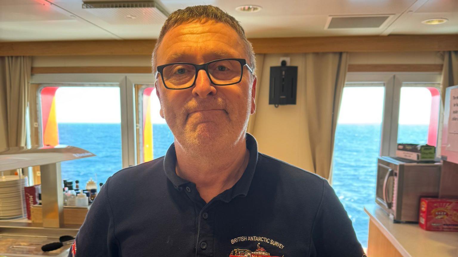 Nick Greenwood is looking at the camera and has short fair hair and is wearing spectacles. He has a black polo-shirt on with the insignia of the British Antarctic Survey ship on the chest. He is standing in a canteen area on a boat. Food can be seen to one side and the blue sea is in the background