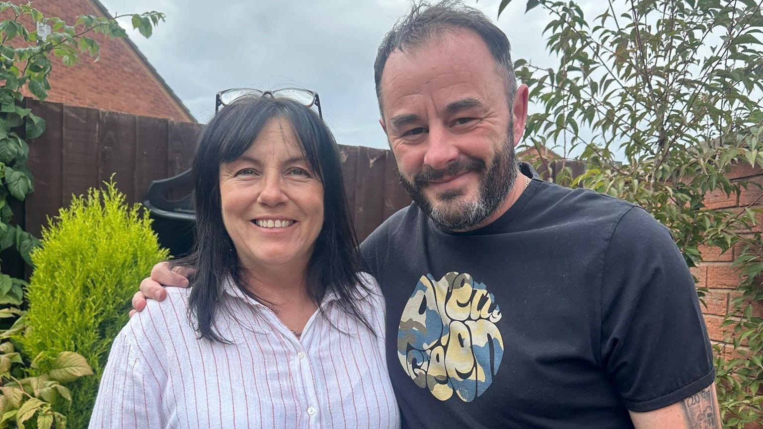 A dark-haired woman in a white and pink striped shirt and a man with his arm around her wearing a black T-shirt