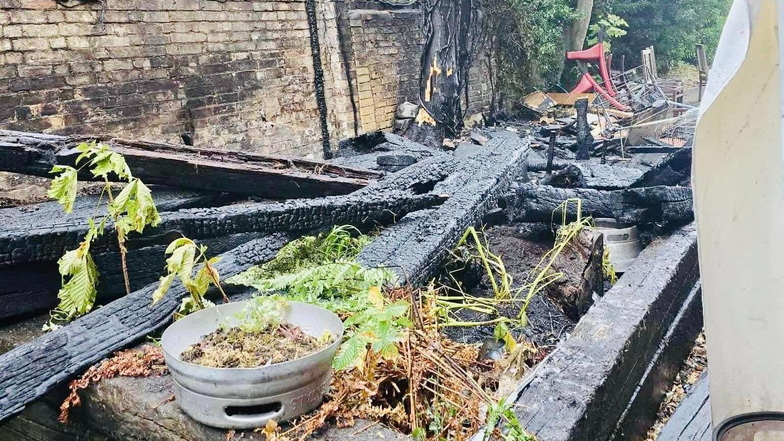 A pile of wood which has been burnt by a fire is standing on top of each other. A brown bricked wall goes along the left side of the image.