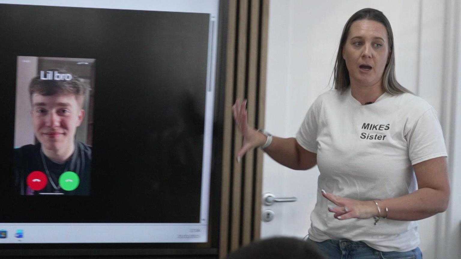 Becky Dustan, wearing a white T-shirt with "Mike's Sister" written on it talks to a classroom of pupils while giving a presentation about the effects of knife crime. On the interactive whiteboard is a screen shot from a phone showing a call coming in from someone called "Lil bro" with a picture of a young man showing.