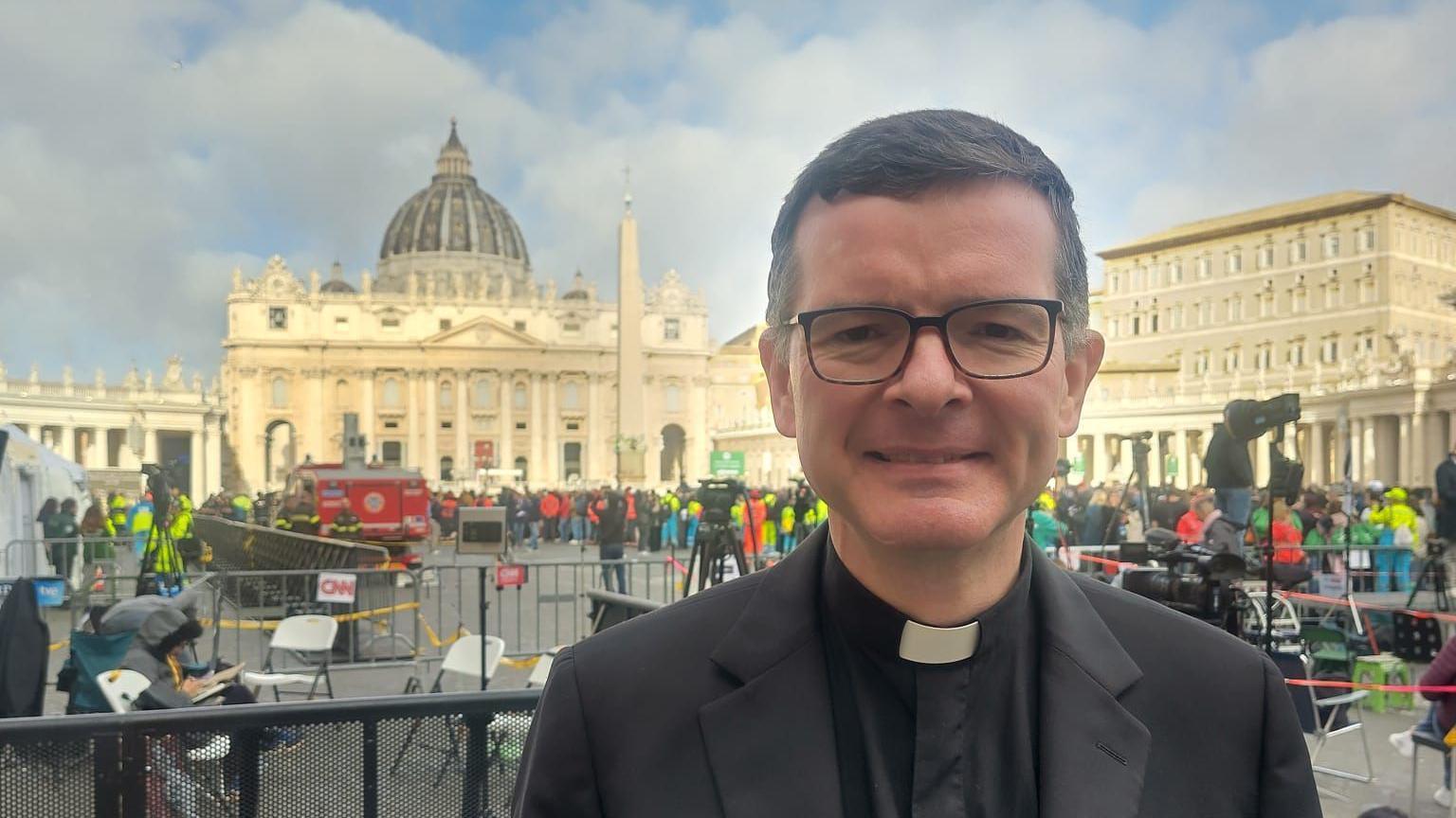 Enda Murphy, who has short, dark hair and wears black glasses and a black suit over black shirt with a clerical collar. Behind him is St Peter's Basilica, a light-coloured stone building with ornate pillars and a grand, domed roof.