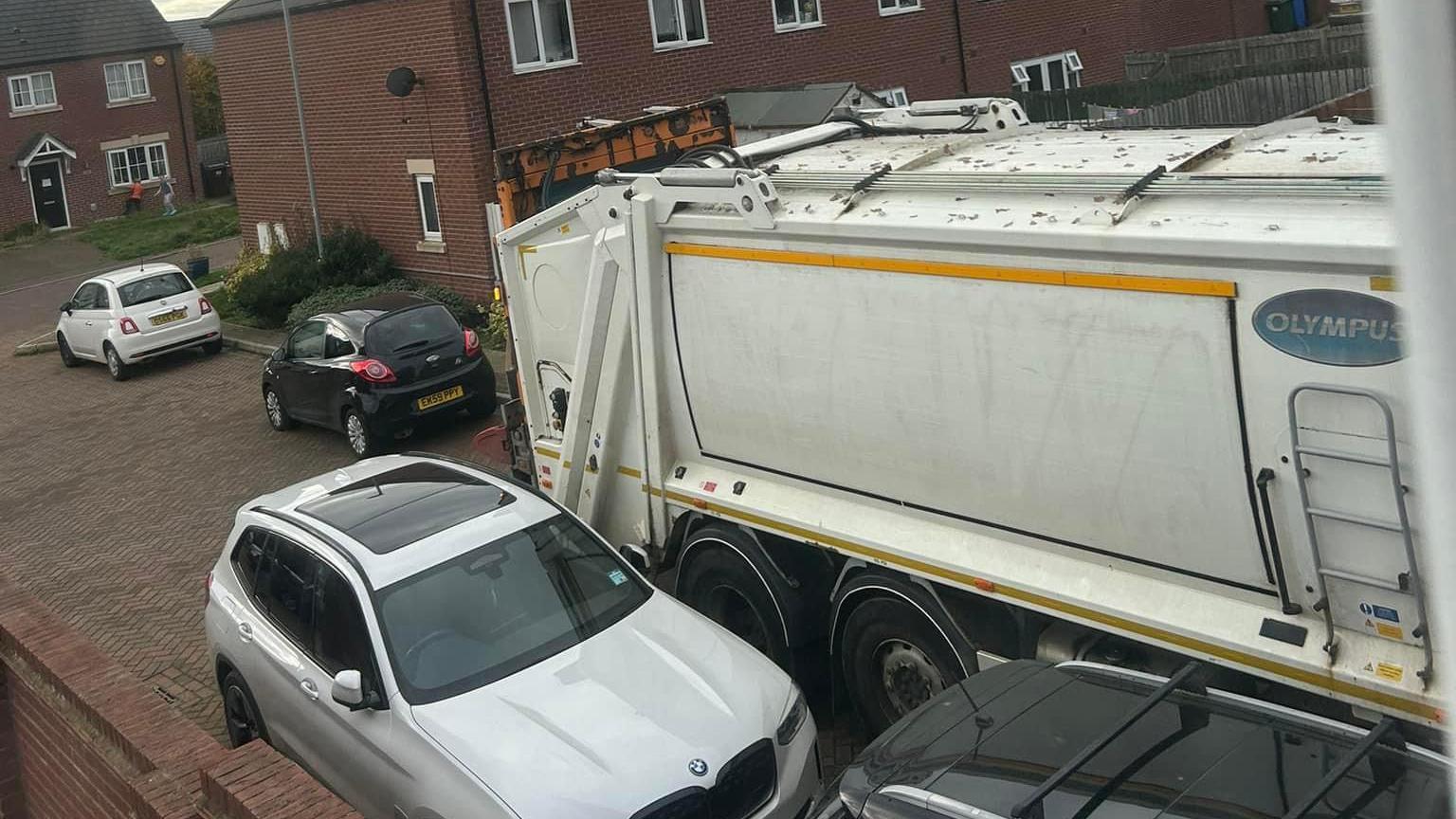 A white bin lorry struggles to pass parked cars in in a residential street in Cottingham. The paved street is part of a new-build housing estate.