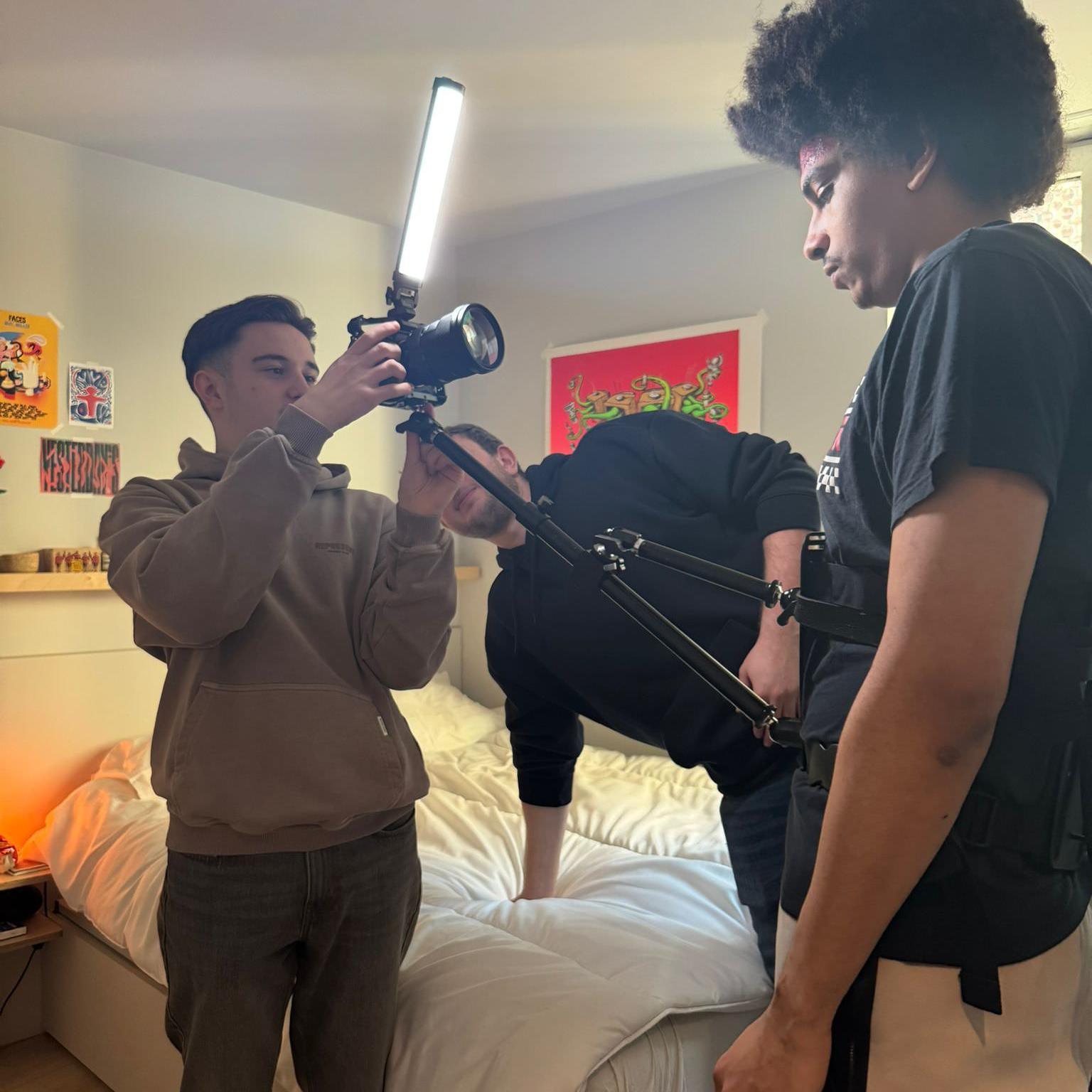Three young men setting up a camera with an LED light in a bedroom, with one person wearing a chest-mounted camera stabilizer and others adjusting the equipment, surrounded by colorful posters and a neatly made bed.