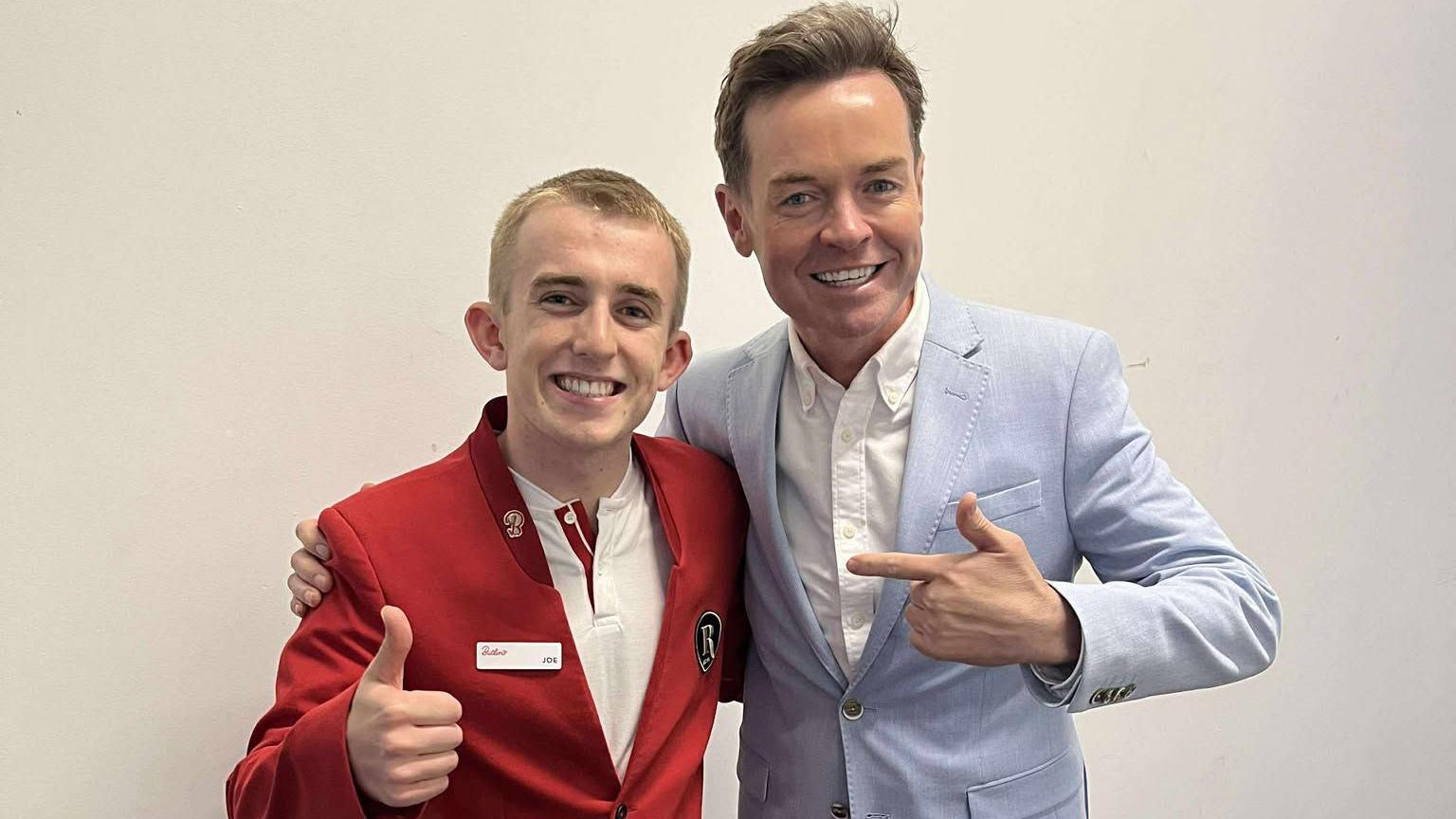 A young man with cropped blond hair and wearing a red jacket and white shirt gives a thumbs up as he stands arm-in-arm with TV presenter Stephen Mulhern, who has short brown hair, is wearing a light-blue jacket and white shirt, and is pointing at the young man.