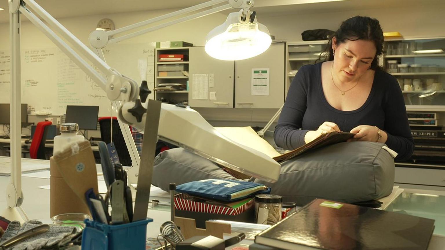 A woman in an office looking through an old ledger with an overhead light over it.
