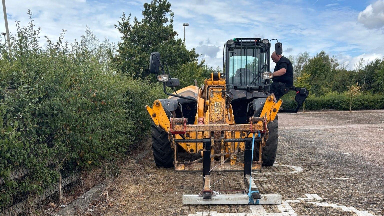 A man exiting the cab of a yellow piece of machinery, which is parked on paved car park stones beside a tall hedge.