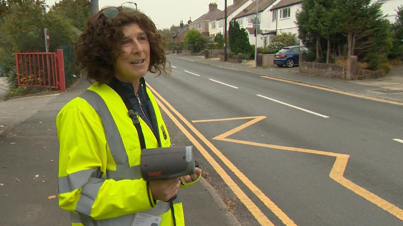 Jane has mid-length curly hair and is wearing a high visibility yellow jacket. She is standing on the side of a road outside a school, next to yellow zig zag road markings and is holding a speed gun. There are houses in the background.