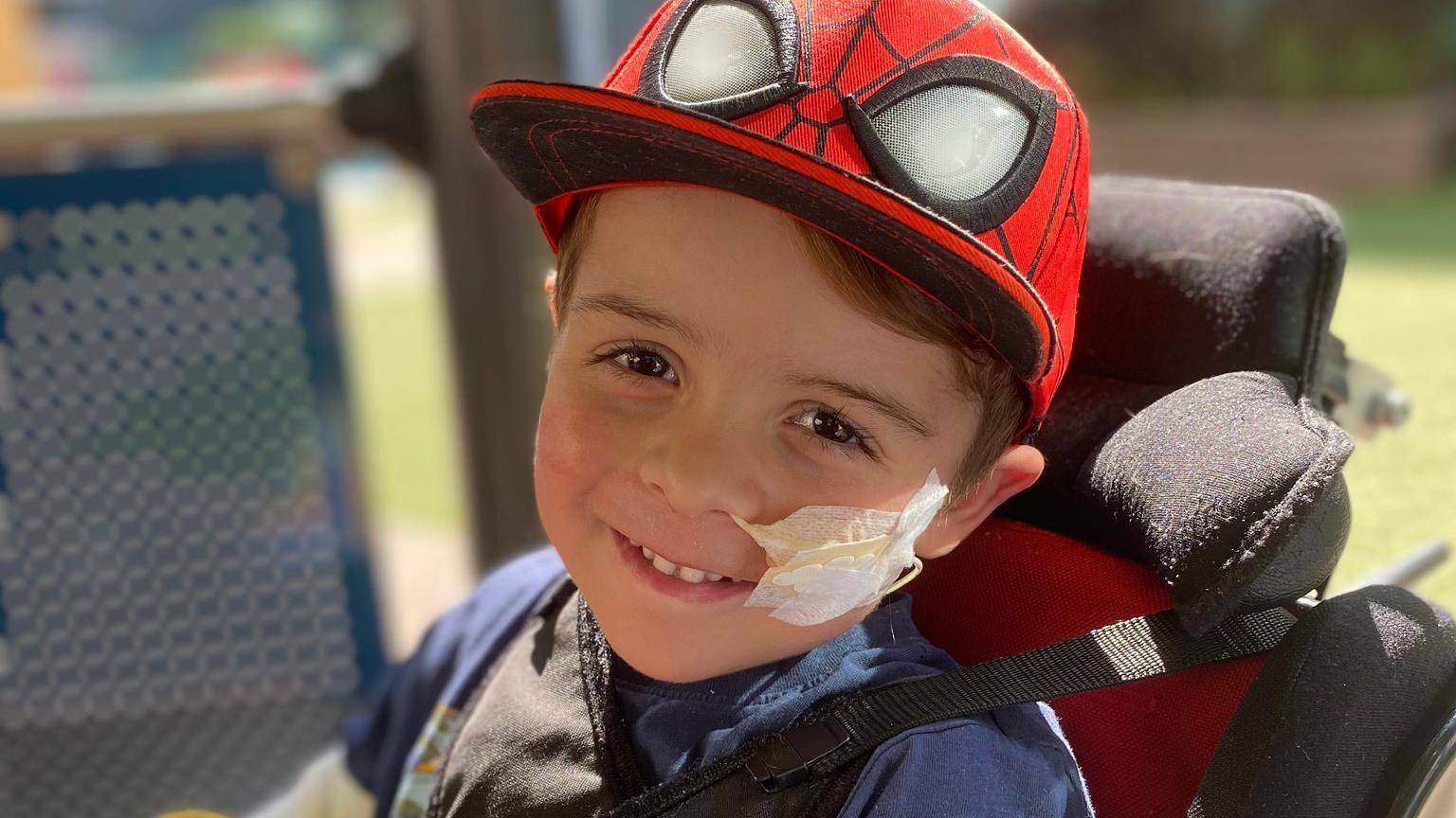 Freddie grinning at the camera, he is wearing a Spider-man cap and is strapped into his wheelchair. He is outside, grass is visible in the background. A tube and tape is on his cheek