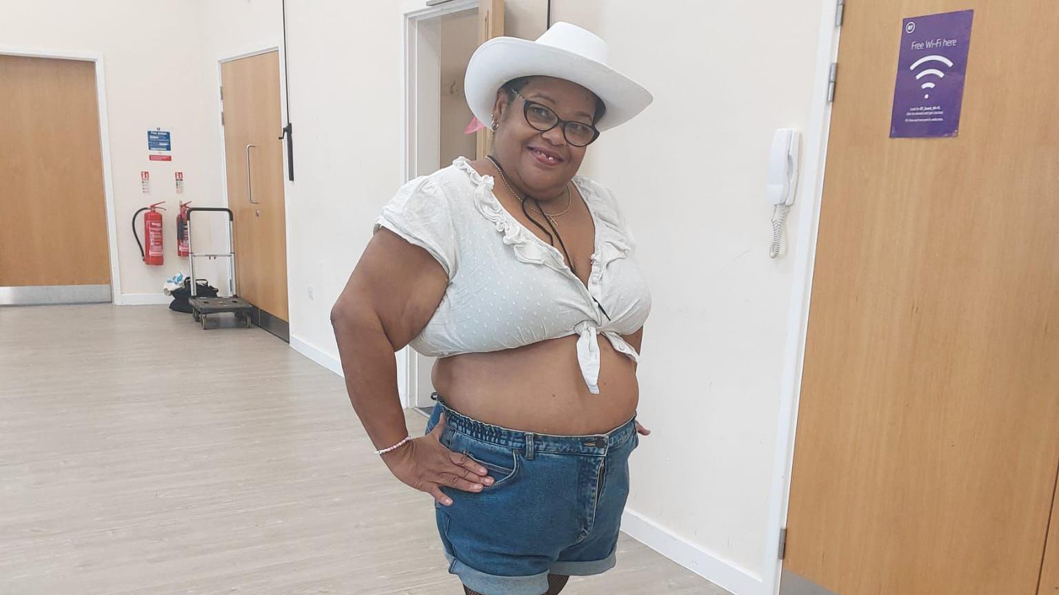 Patsy Wright with short dark hair and glasses wearing blue denim shorts, a white top and a white cowboy hat. Her hands are on her hips. She is standing on a light-coloured laminate floor in a corridor where three wooden doors are visible.