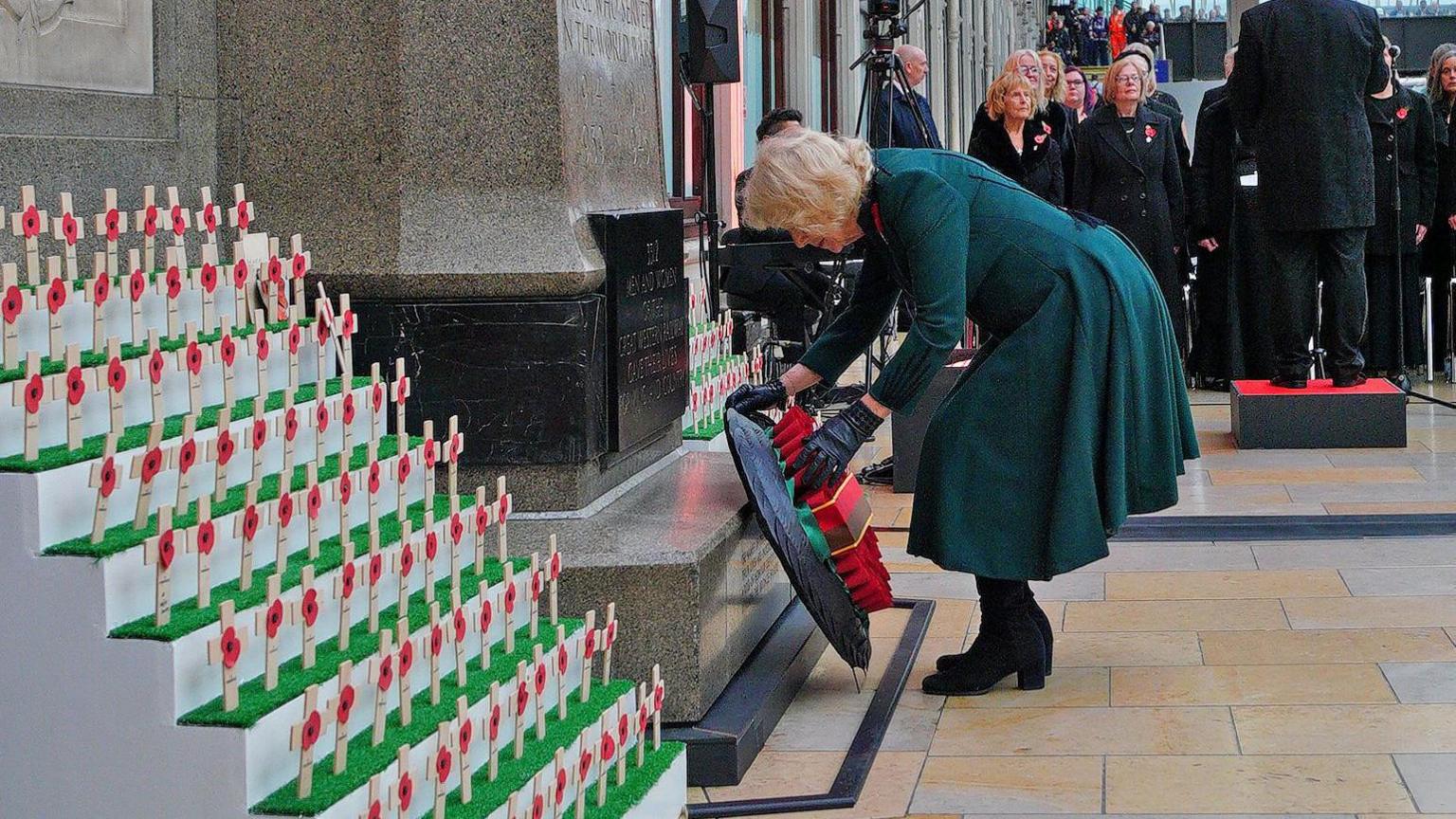 Queen Camilla bends down to place her wreath at a memorial in Paddington station. It is surrounded by a display of small wooden crosses with poppies on. Behind her, a choir is singing.