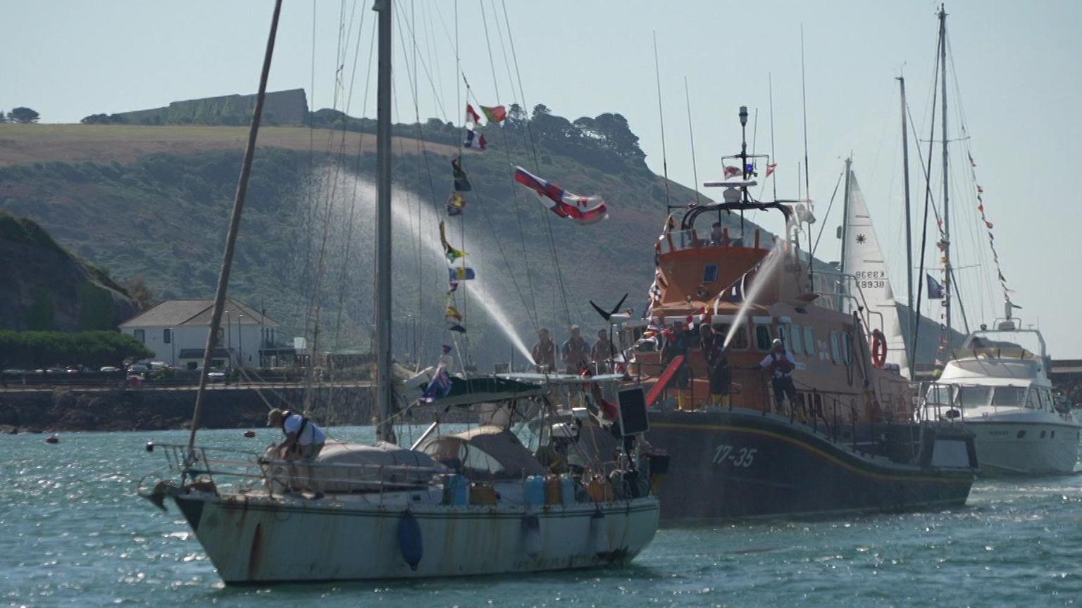 This picture shows a small sailing boat with a person aboard.
Just behind it, an orange RNLI rescue vessel is spraying water toward the sailboat.
The background features several other boats and yachts, with a coastline of hills and buildings.