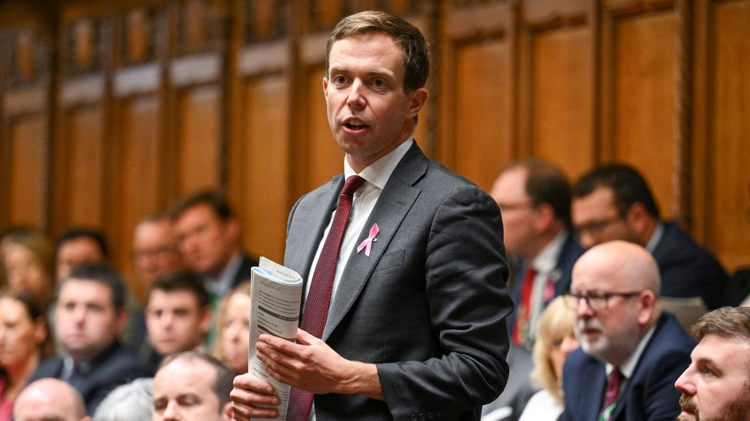 A man, wearing a grey suit stands among a group of people, mainly men, in the House of Commons. He is holding a piece of paper on his hands.