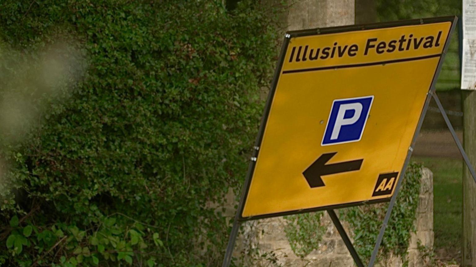 A yellow temporary road sign saying Illusive Festival with an arrow and a white letter P in a blue rectangle against a hedge