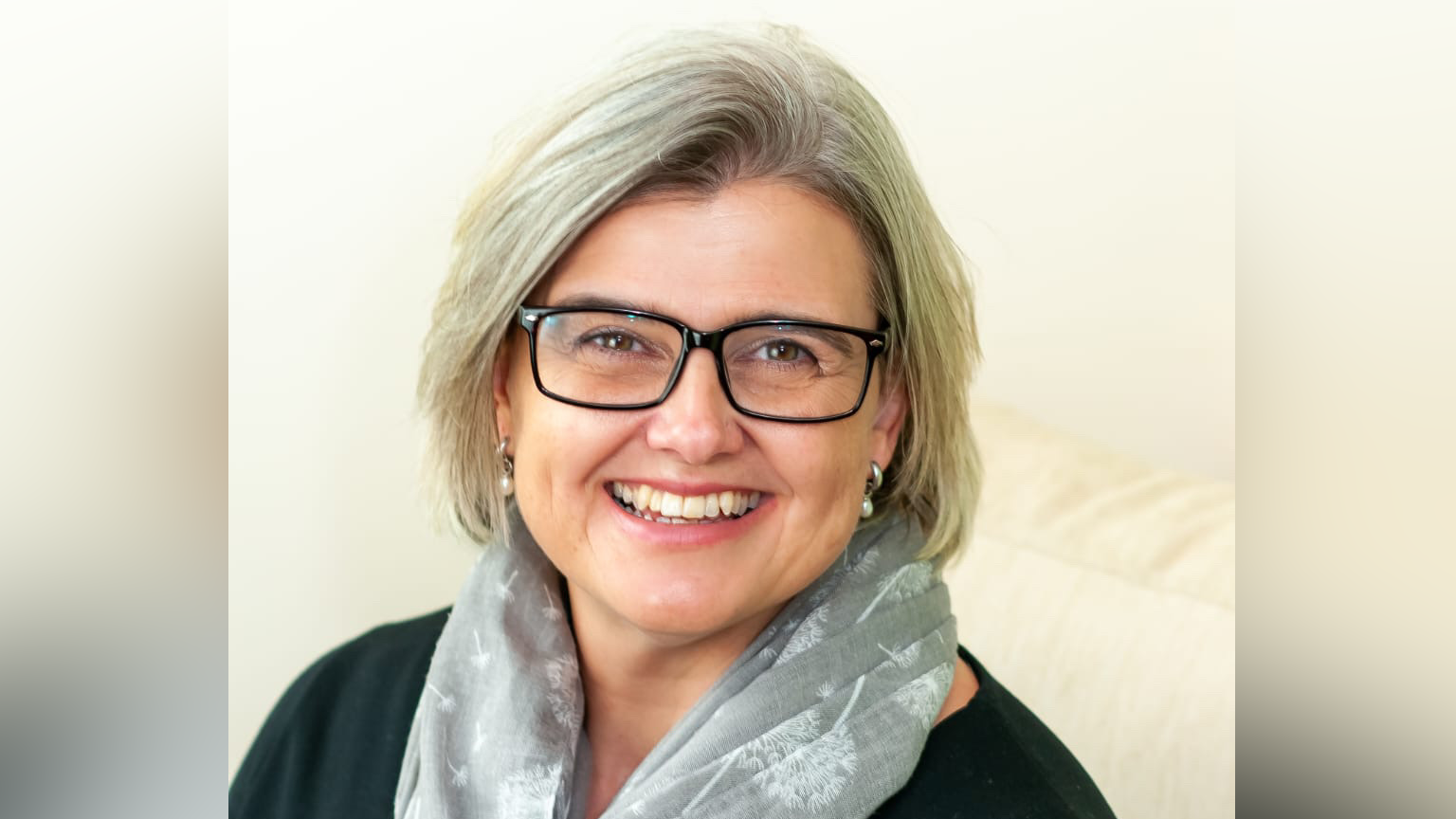A head shot image of Ms Whymark. She smiles over her shoulder toward the camera. She has a blonde bob hairstyle and wears black glasses, pearl earrings, a black top and a light green shirt with some white floral details on it.