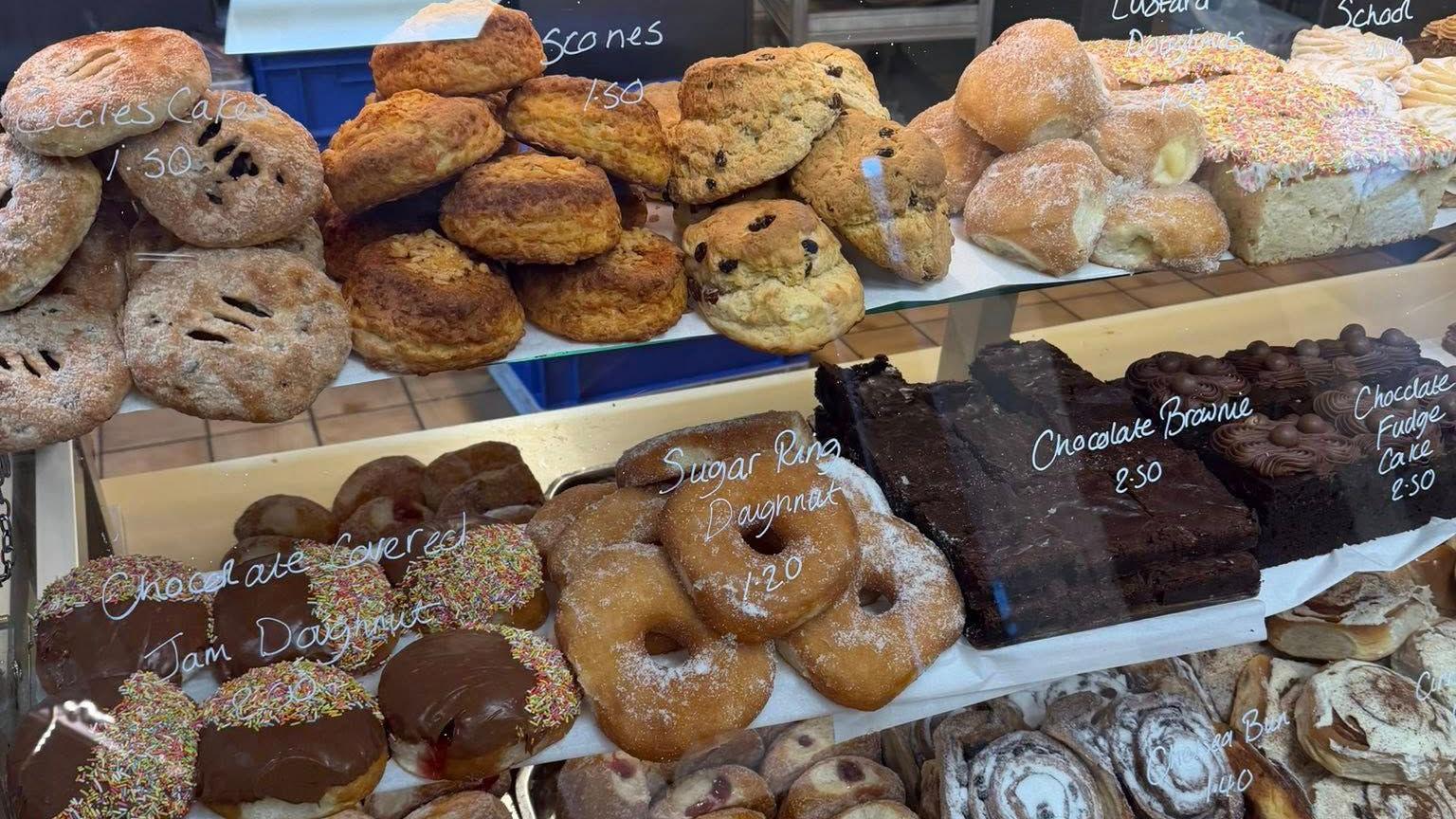 Baked goods behind a glass cabinet, including chocolate jam doughnuts, scones, Eccles cakes and chocolate brownies.