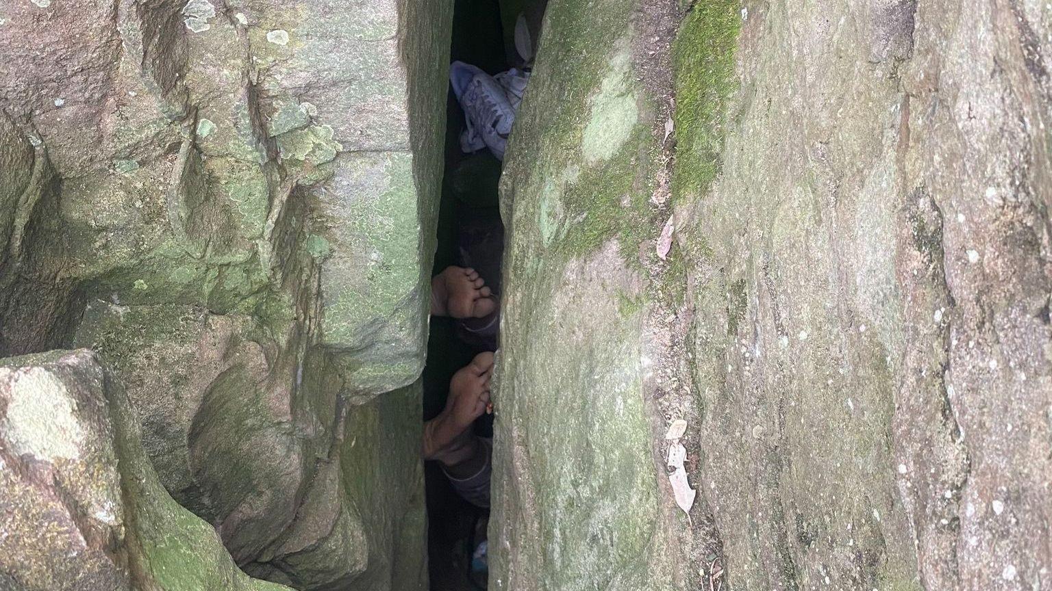 The bottoms of two feet visible between two large boulders. A pair of shoes can be seen to the side inside the crevice