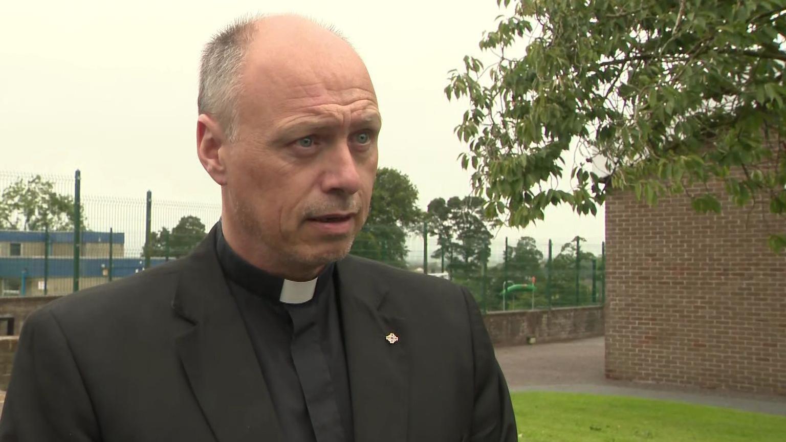 Fr Martin Graham is standing next to a brick building, with a tree on the right. He is wearing his dog collar and black priest attire. He has thinning hair and blue eyes.