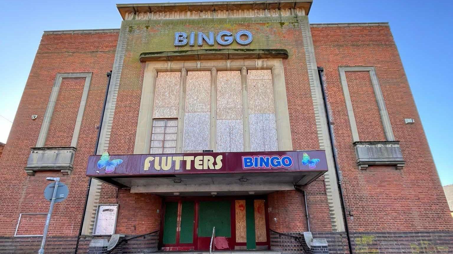 The outside of a large art deco brick building, with large windows, that have been boarded up. It has an entrance, also boarded up, a sign, and steps leading to the front. 