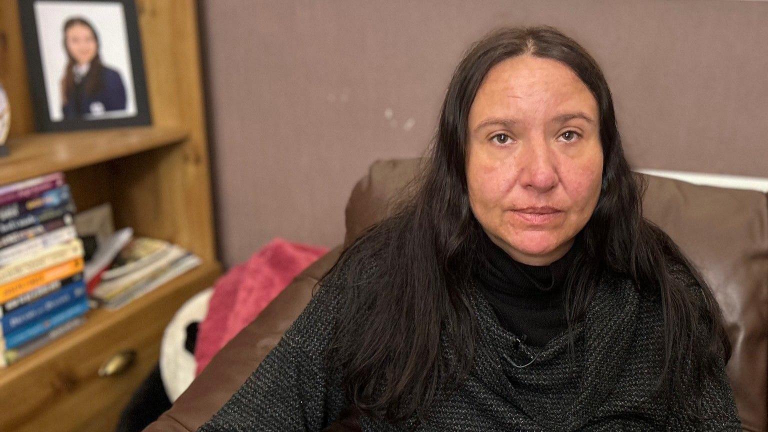 A woman is seated on a sofa, looking at the camera. She has long, dark hair and is wearing dark clothes. Behind her, slightly out of focus, is a portrait of her late daughter. 