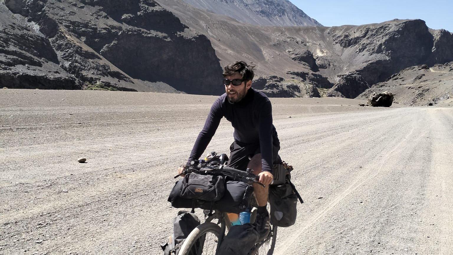Man with dark hair and sunglasses cycles along a deserted track with mountains behind
