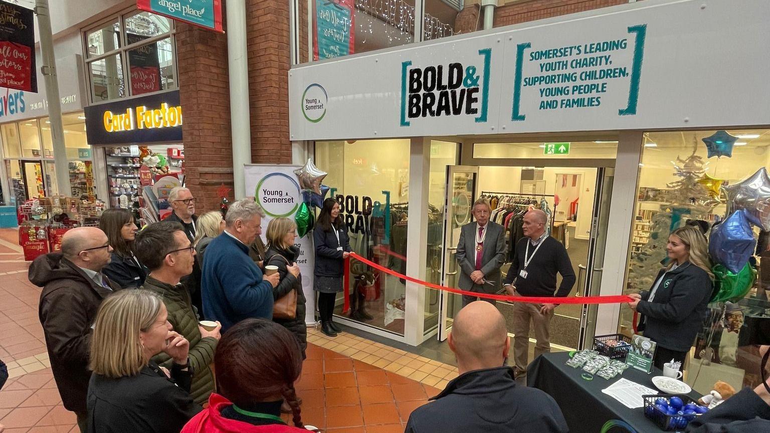 A row of people standing front of a red ribbon waiting to see it get cut in half in front of a windowed shop called Bold and Brave. There is a Card Factory shop next door and a Savers next to that.