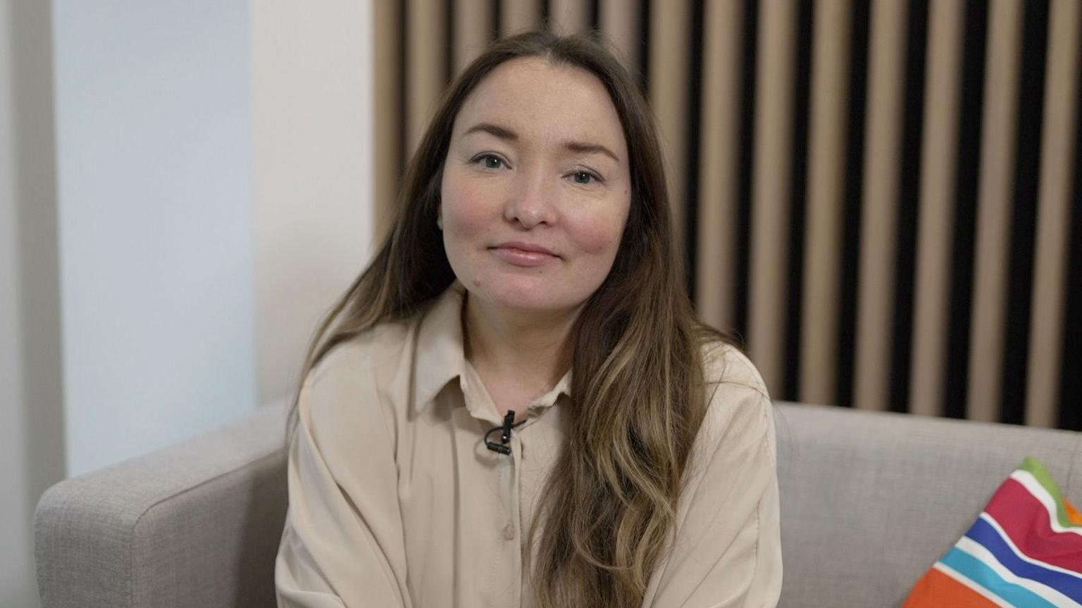 Dr Elvira Bolat, Associate Professor in Digital Marketing, sat at Bournemouth University.  She is sitting on a grey sofa with a colourful cushion. Her long brown hair is over her shoulders and she is wearing a beige shirt.