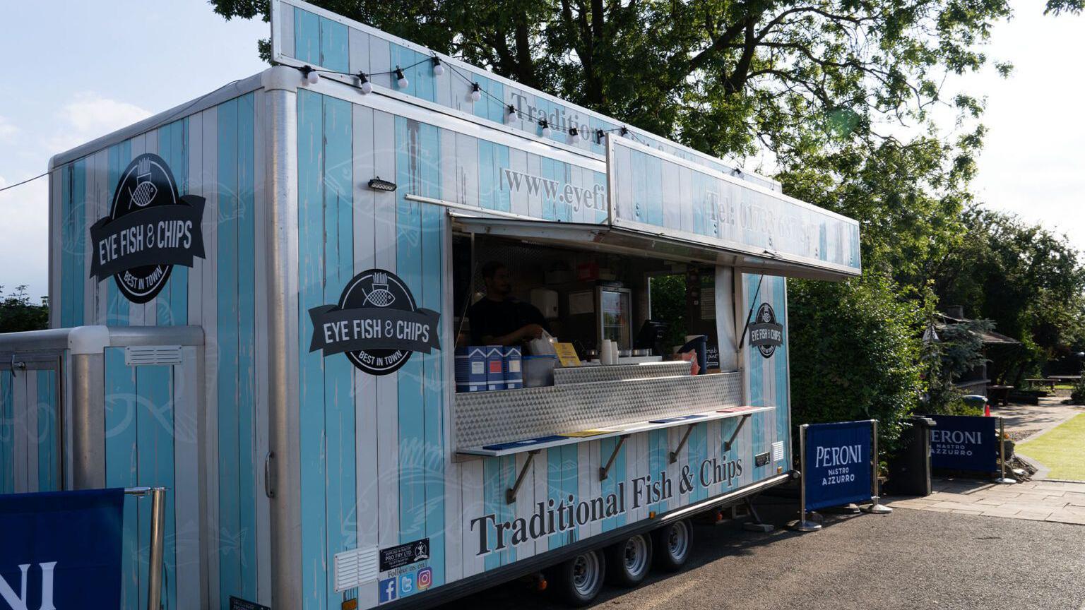 A blue and white fish and chip trailer next to a beer garden of a pub. Signs on it say Eye fish and chips.