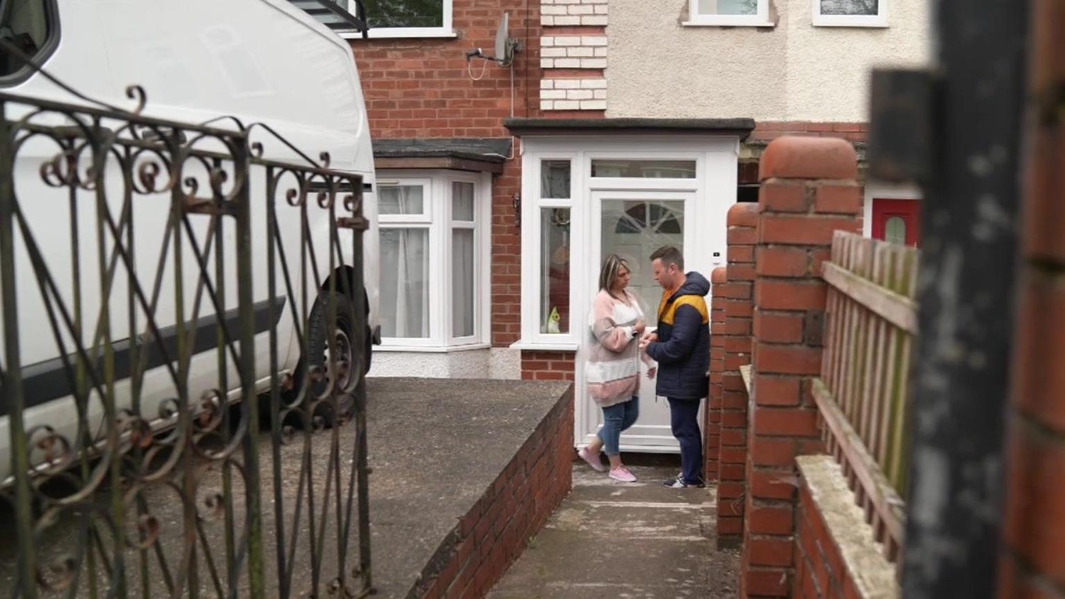 Rebecca and Peter outside the front door of their house, which has a white porch and a brick and wooden fence along the path to the door