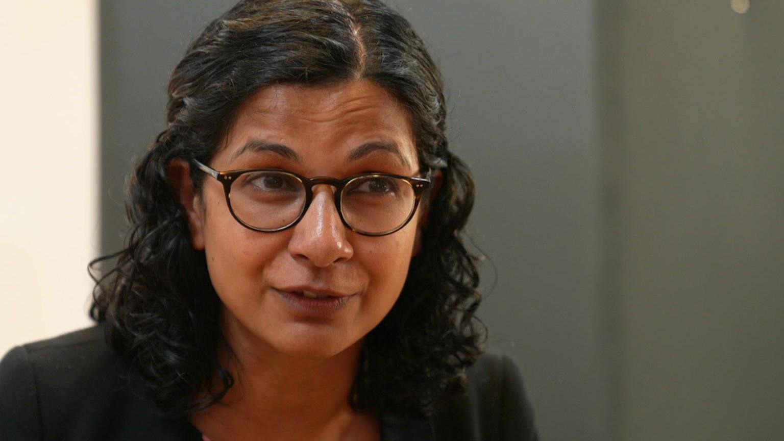 A woman with curly dark hair and black rimmed glasses. She wears a black blazer and a polka dot dress or top. She looks to be of South Asian heritage.