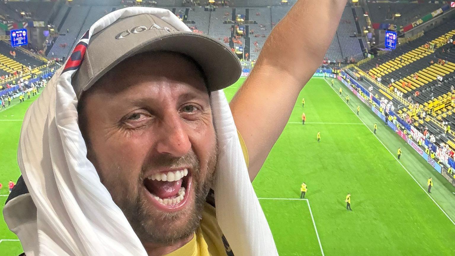 Alex Fox looking into the camera smiling in front of the pitch wearing a cap