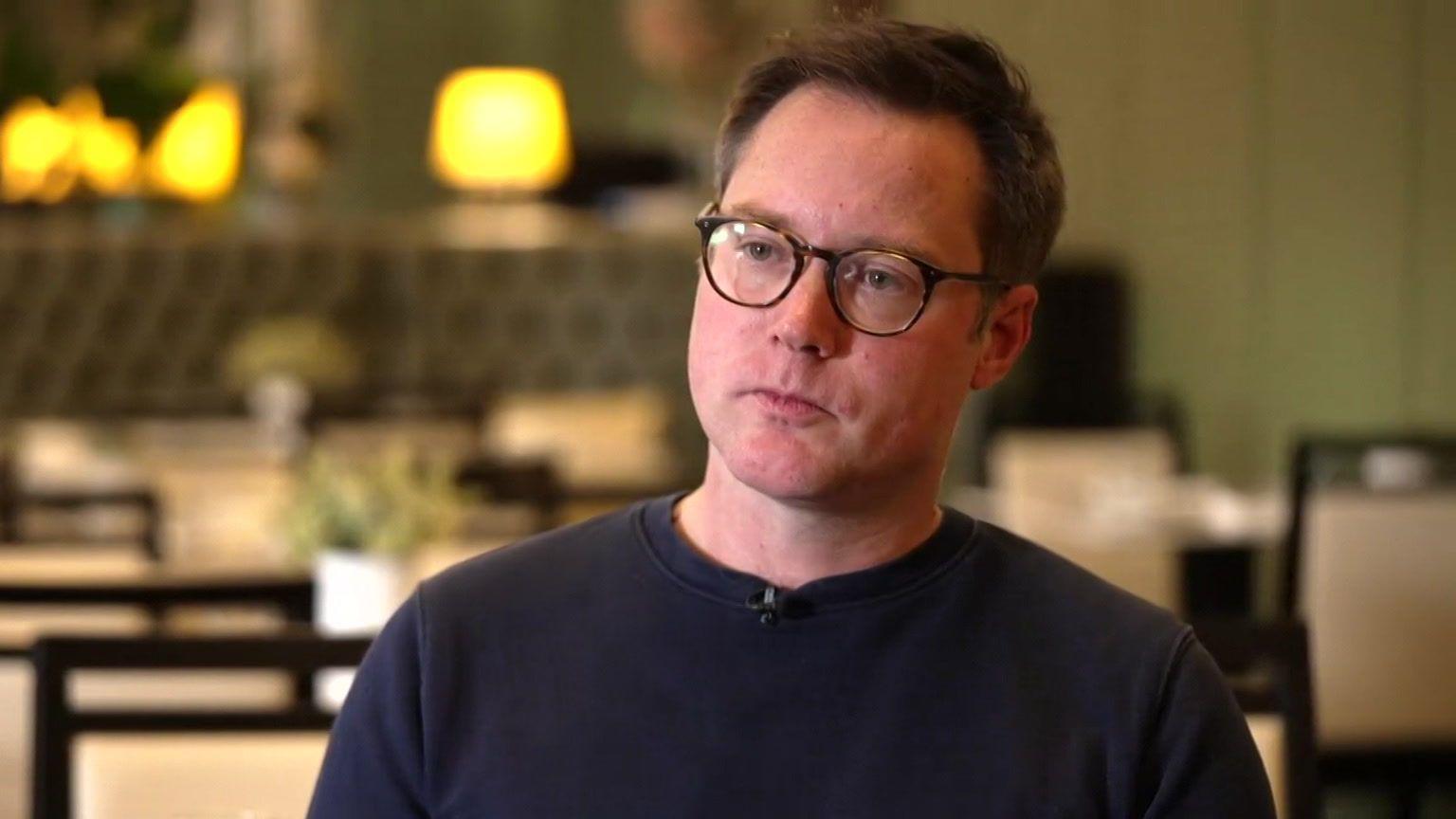 Allen Simpson is seen sitting in a restaurant during an interview. Tables and chairs are visible in the background