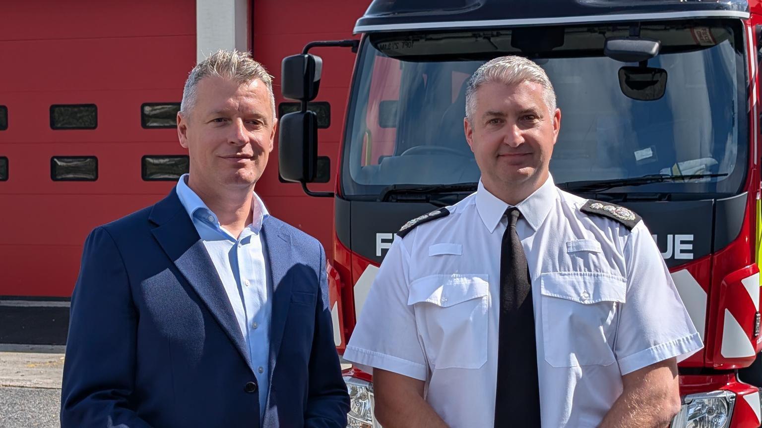Luke Pollard and Gavin Ellis stood in front of a fire engine parked in front of the fire station. Pollard is wearing a blue shirt and blue suit jacket. Ellis is wearing a white shirt with a black tie. They are both smiling at the camera.