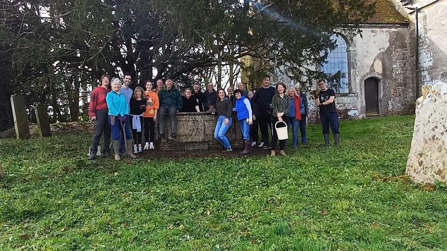 A large group of people gather in the churchyard around a concrete tomb, there are graves in the background as well as trees and a section of the church can also be seen