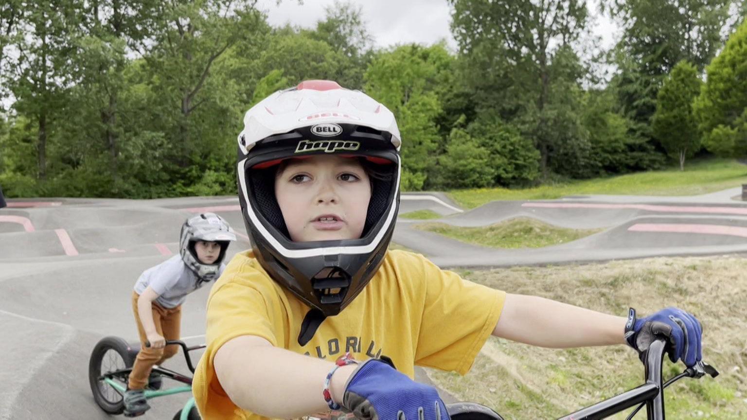 Ten-year-old Tom sitting on his bike during an interview. He is wearing a white full-face helmet and purple riding gloves. His seven-year-old brother Felix is riding onto the track on his BMX behind him.