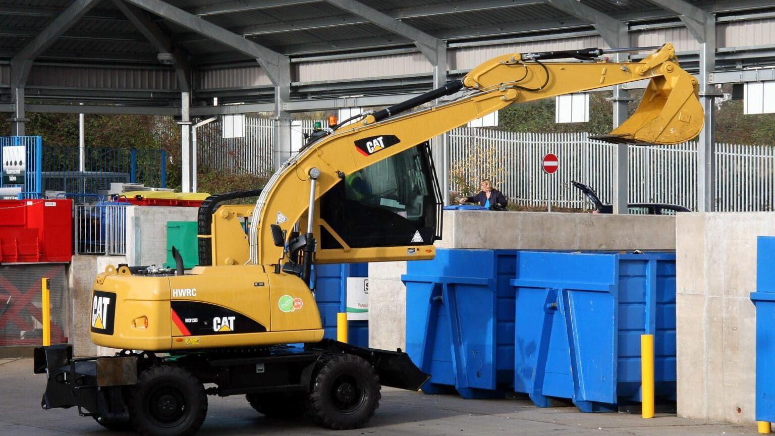 A yellow front loader tractor being driven by a person. The scoop is above two blue bins. 