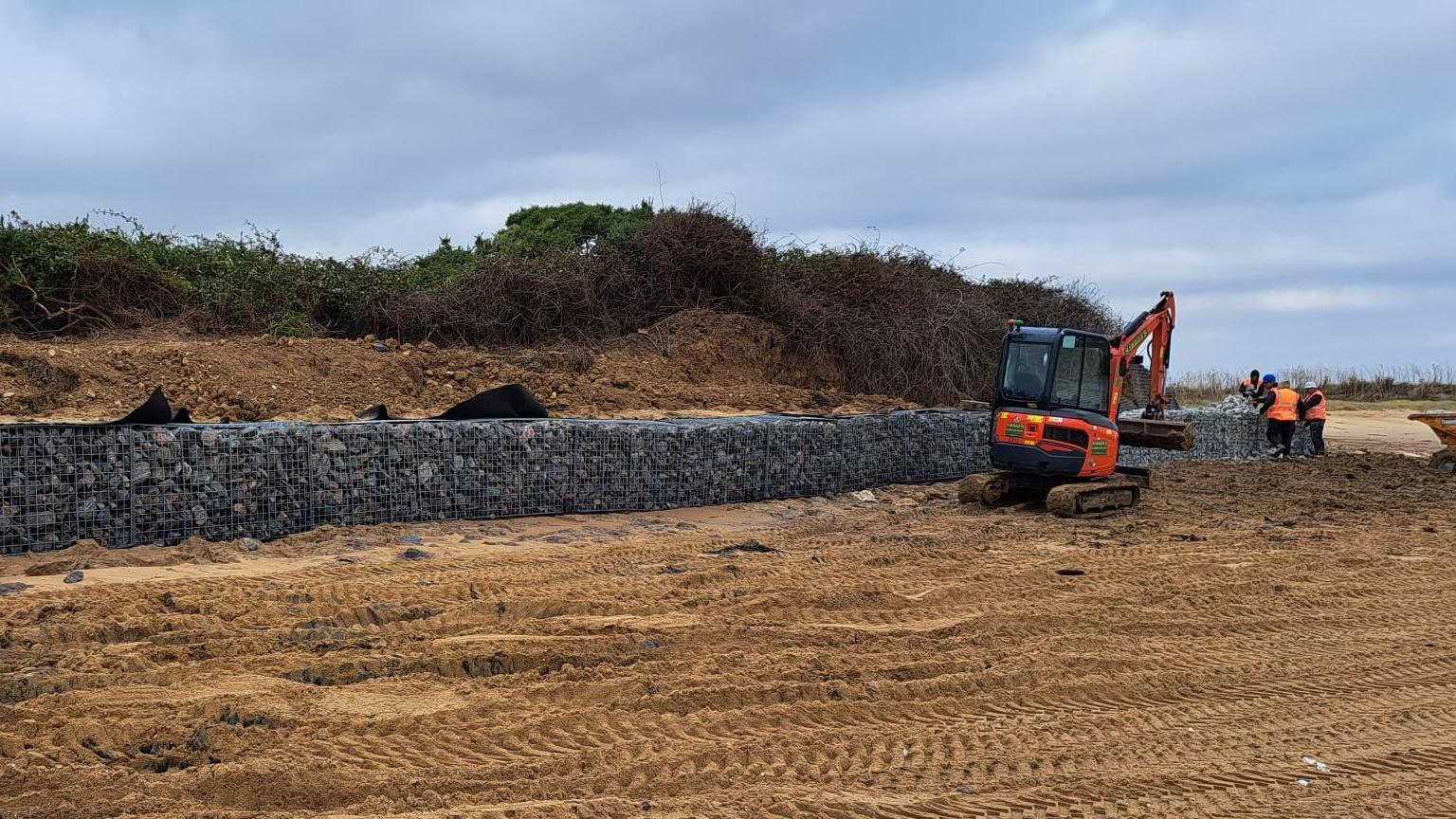 A digger and workers on the beach at Walton-on-the-Naze. There is a long sea wall built of stones on the sand