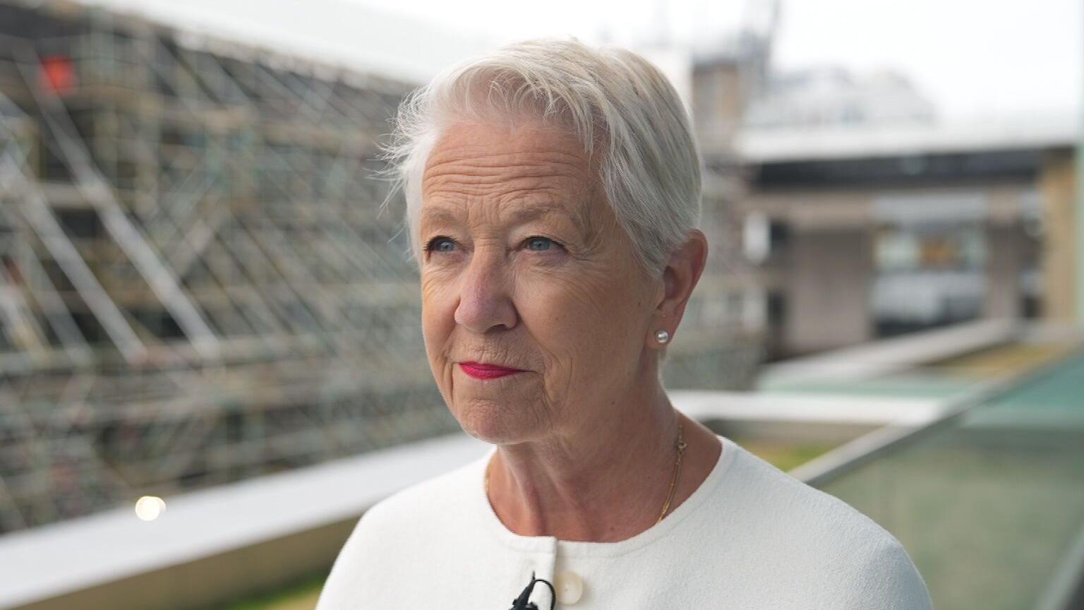Ann Priest - a woman wearing a white outfit, with white hair, standing outside, and looking off to the left slightly.