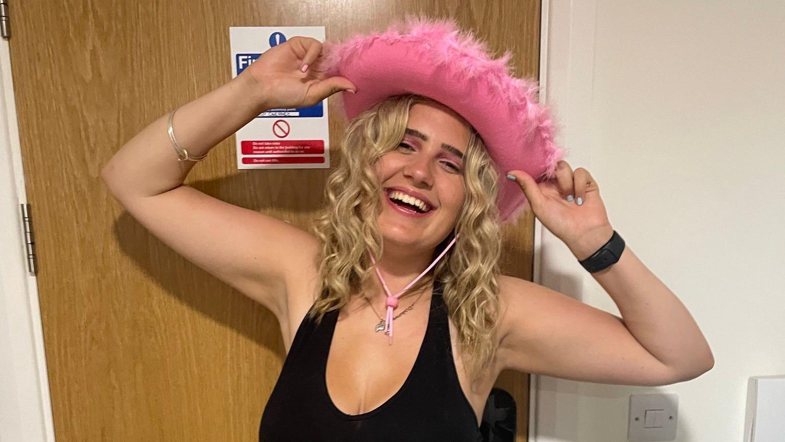 Phoebe who has mid length blonde curly hair is posing in front of a light brown wooden door. She is smiling whilst wearing a pink cowboy hat with a feathered trim. She is holding the cowboy hat on her head with both hands and her arms raised.