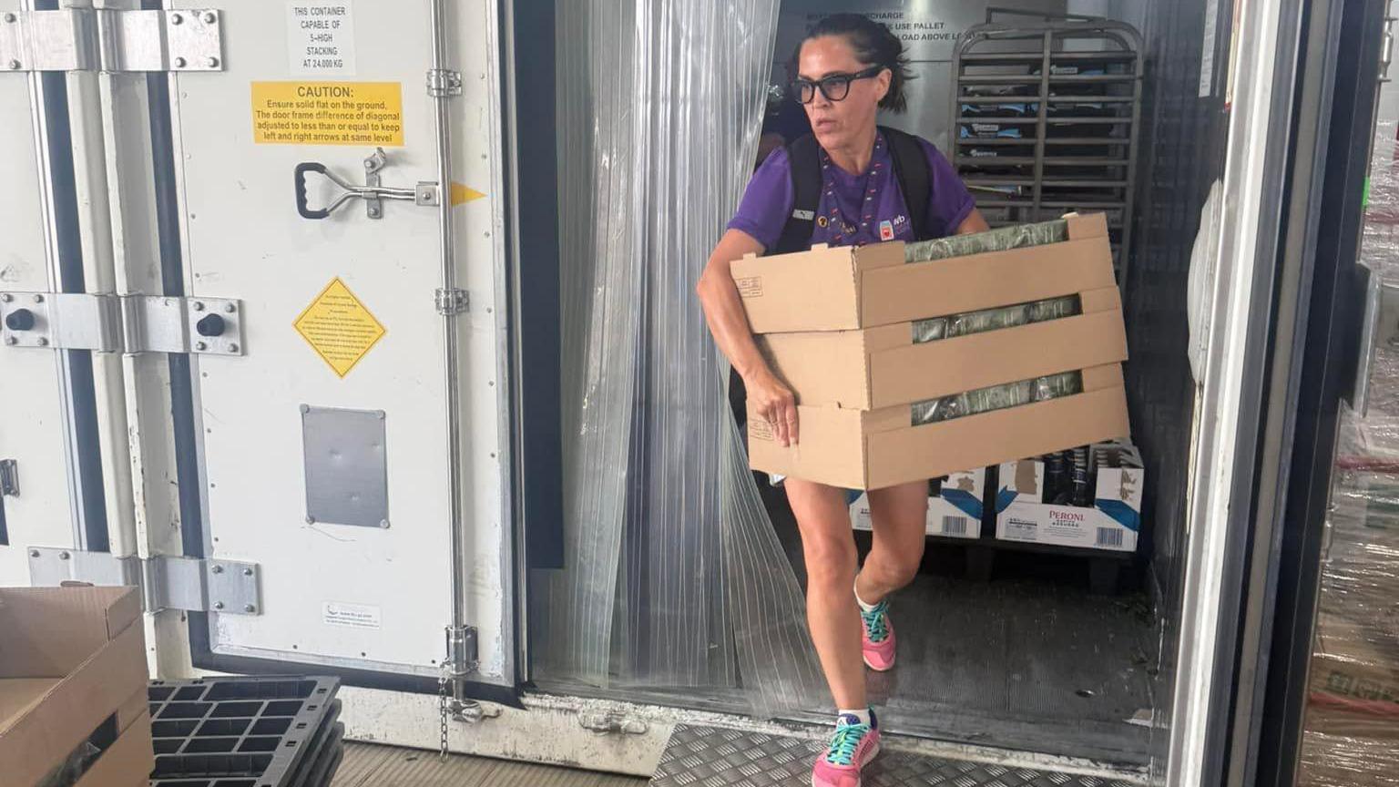 A woman, wearing shorts and a purple T-shirt, with a backpack on her back, walks out of a cold storage unit holding three boxes of food. She has dark hair tied back and is wearing glasses.