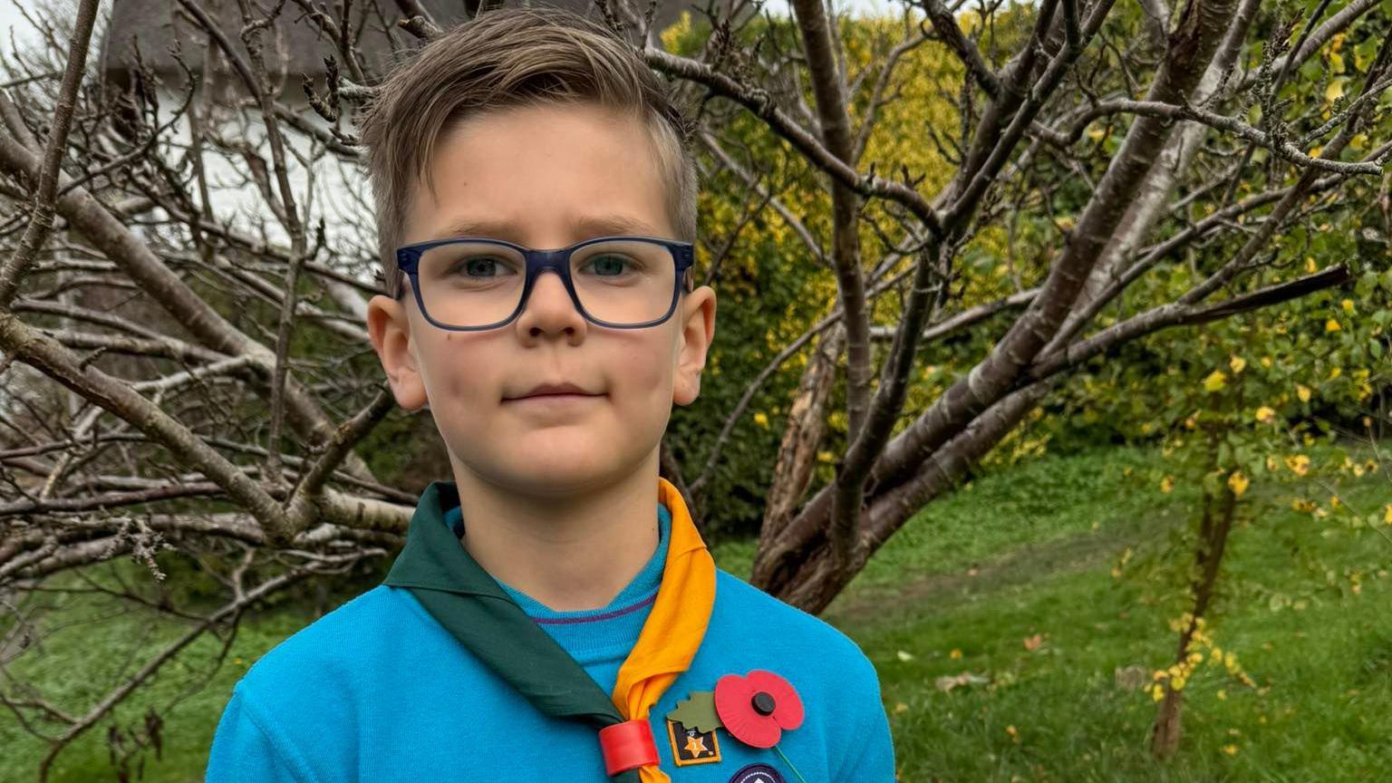 Boy with brown hair and glasses wearing a blue jumper and yellow and green piece of fabric and a poppy.