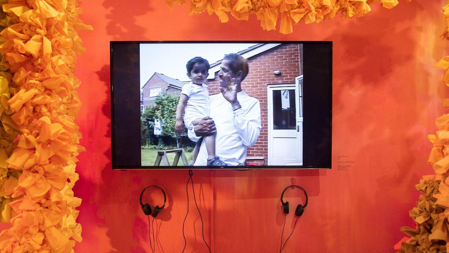 A TV screen displays an old video of an Indian man holding his young daughter in their back garden.