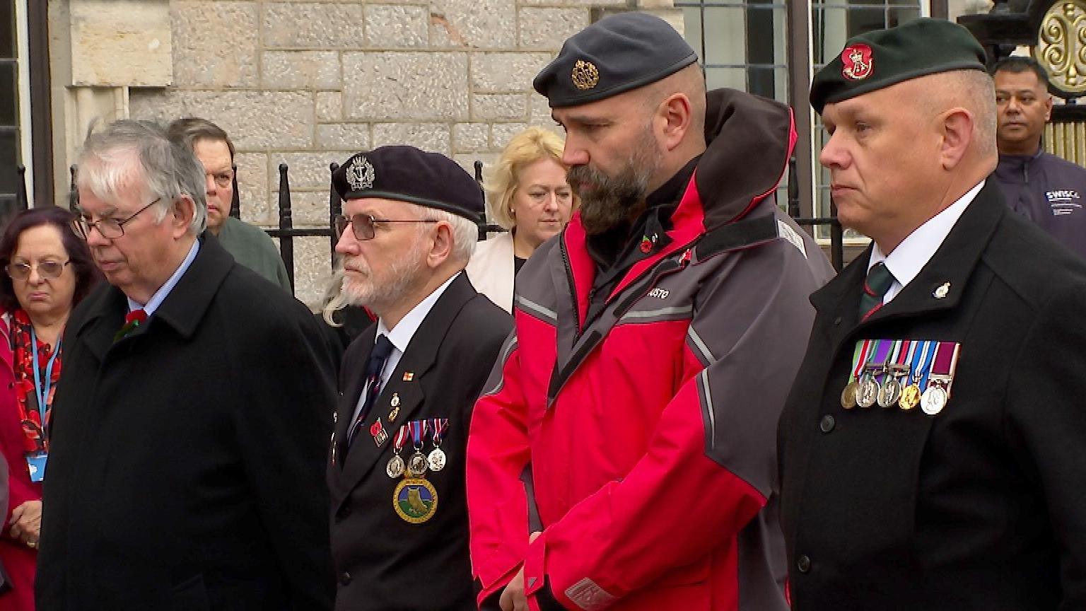 Four men standing with their heads bowed in respect. They are all dressed smartly, with three wearing berets, and the other wearing a black jacket. 