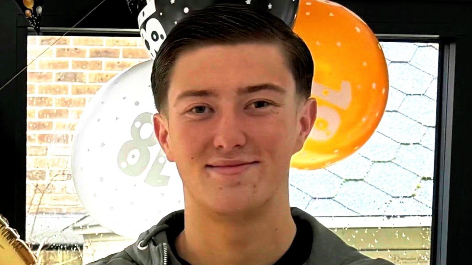 A young man with short dark hair is smiling to camera. Balloons with 18 on them can be seen in the background